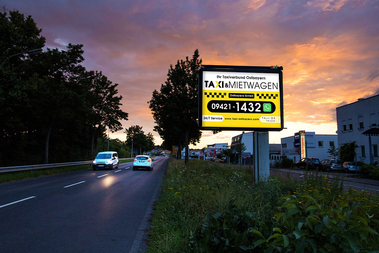 Plakatmotiv des Taxi- und Mietwagenverbunds Ostbayern an einer Straße bei Sonnenuntergang mit Telefonnummer und Webadresse.
