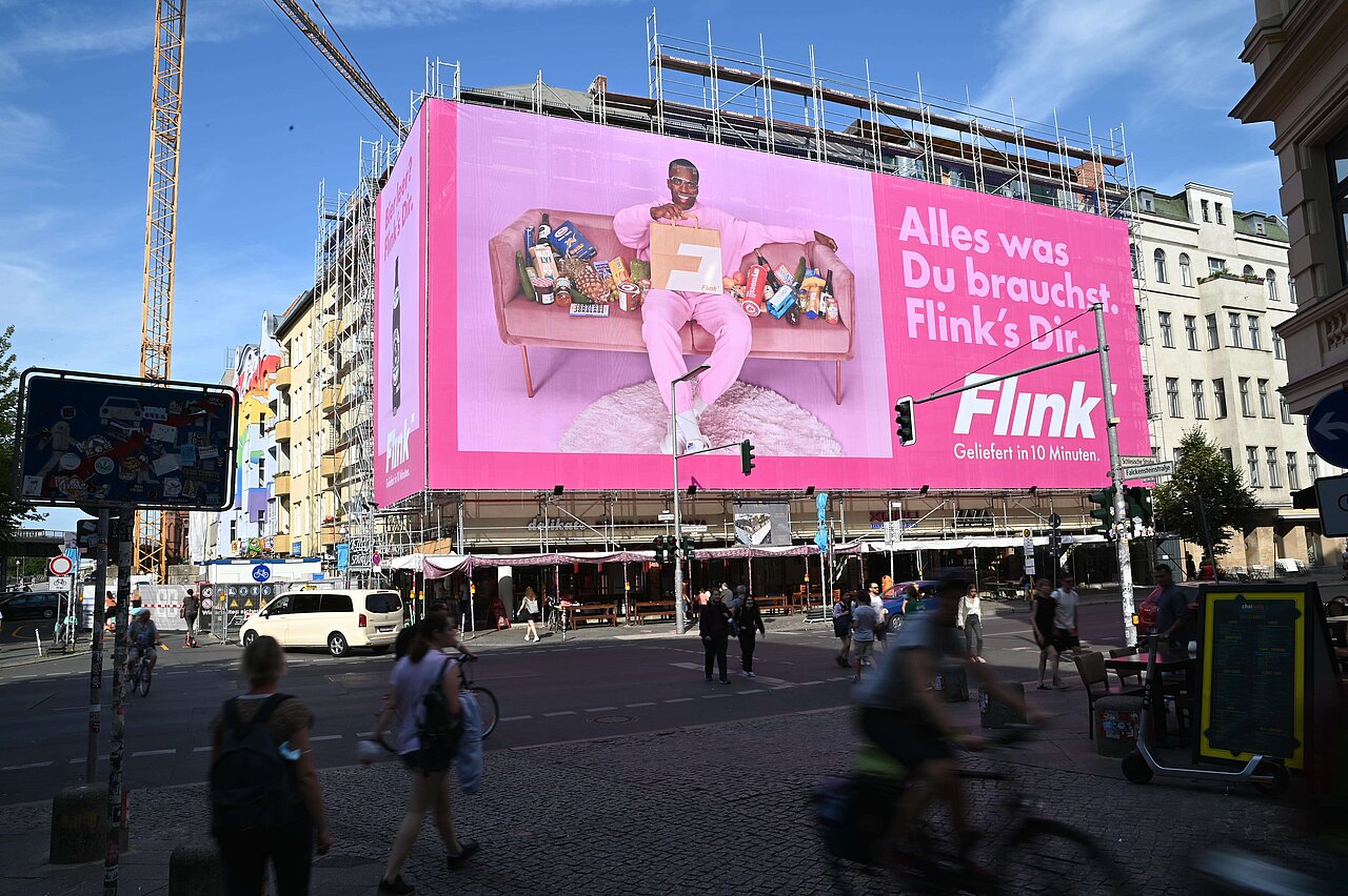 Das XXL-Poster von Flink dominiert die Straßenszene mit einem entspannten Mann in Pink und bewirbt schnelle Lieferungen in nur 10 Minuten.